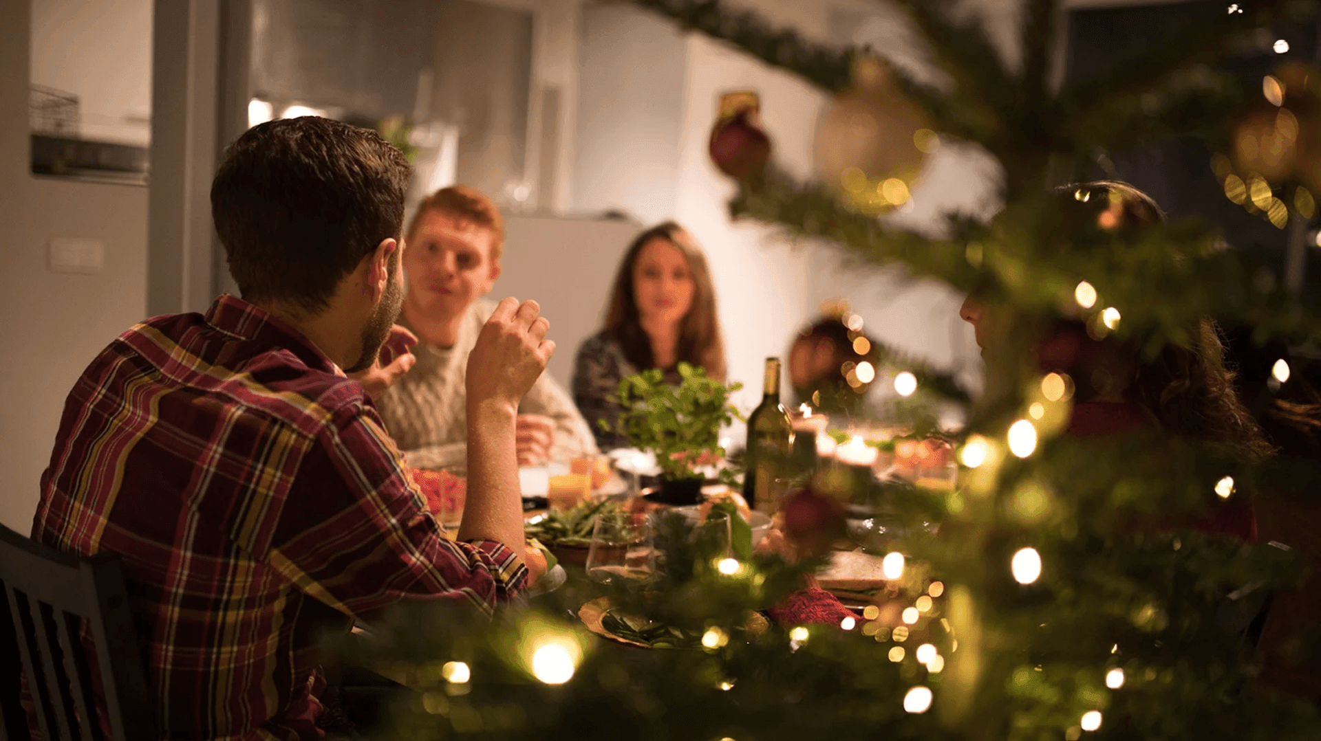People gathered around a dining table, visible through a decorated Christmas tree with lights and ornaments.